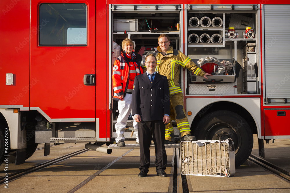 Obraz premium Portrait of happy firefighters and paramedic woman standing together before fire engine truck
