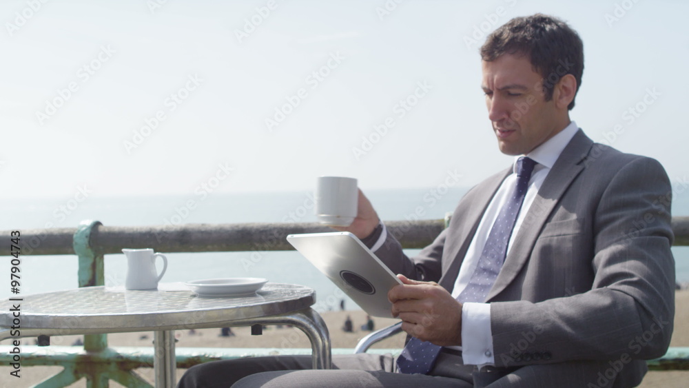Businessman using computer tablet and drinking coffee at seafront cafe. Shot on RED Epic. Businessman using computer tablet and drinking coffee at seafront cafe. Shot on RED Epic.