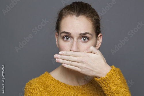 negative feelings concept - portrait of surprised beautiful 20s girl covering her mouth for bad breath or taboo,studio shot on gray background photo
