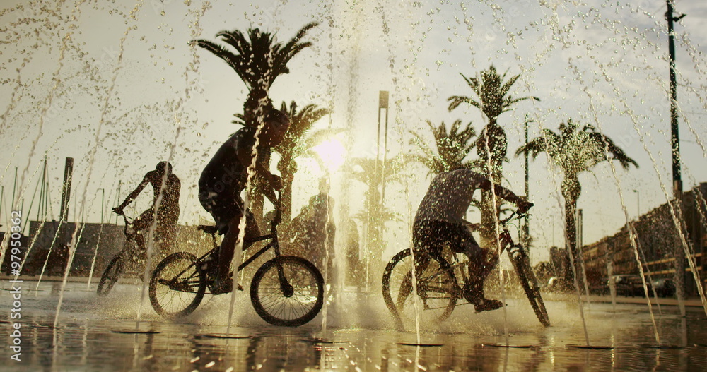  Young boys on bikes cooling off in fountain in the city in summertime. 