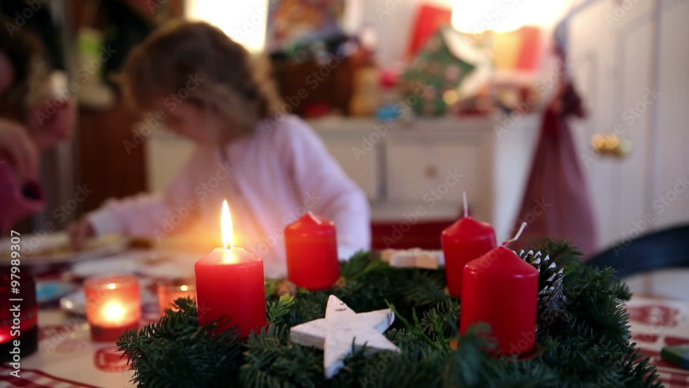 A close up of a burning candle on top of a wreath with a little girl helping her mother making cookies in the background on advent first