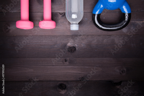 Sport equipment. Blue kettle bell, water bottle and pink dumbbell on a black wooden background