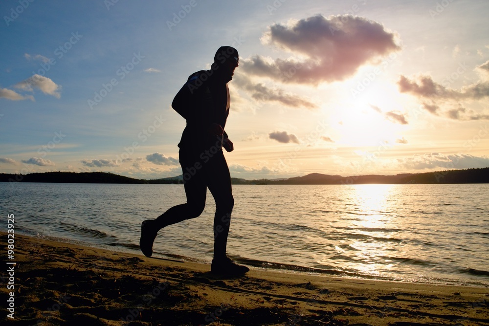 Tall man with sunglass and dark cap is  running on beach at autumn sunset