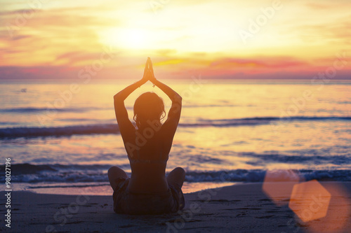 Silhouette of young girl in yoga pose sitting on the beach during sunset. photo