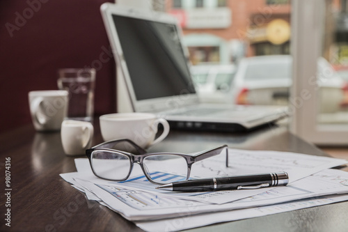 На столе лежат аксессуары для бизнеса: очки, бумаги, ручка, ноут-бук, чашка кофе