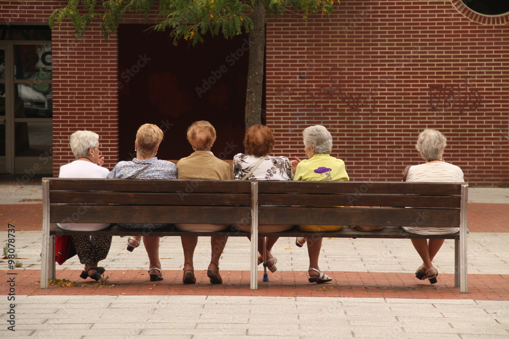 Mujeres jubiladas sentadas en un banco y una de ellas apartada del grupo - obrazy, fototapety, plakaty 
