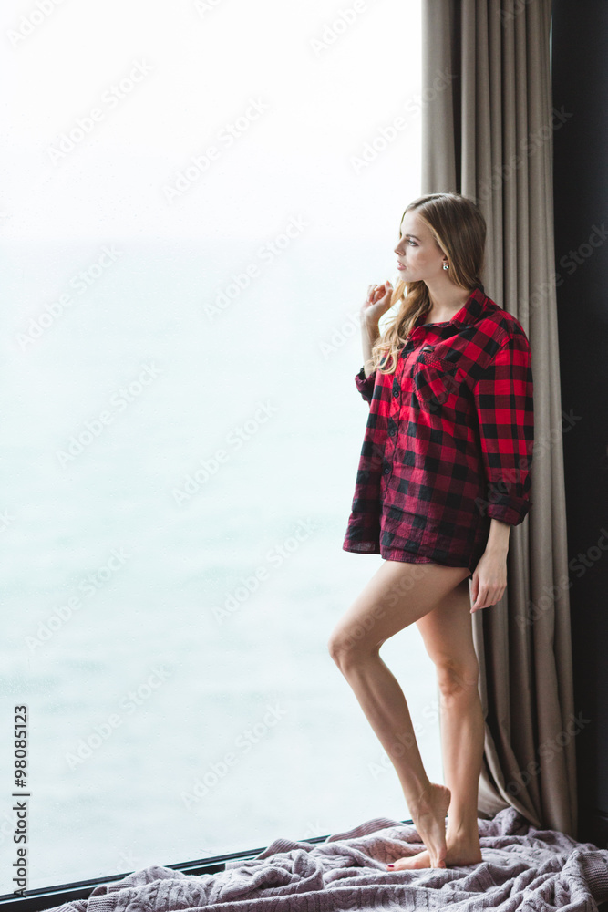 Attractive thoughtful young female standing and looking through the window