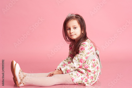 Cute baby girl 4-5 year old wearing trendy floral dress, sitting in room over pink. Looking at camera.