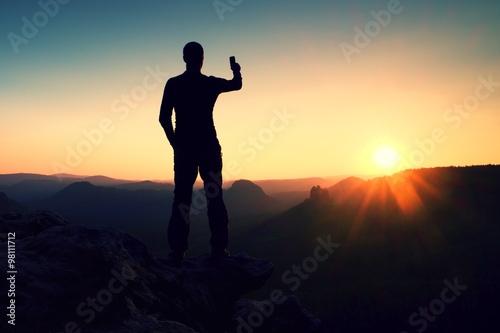 Selfie mania. Tall man hiker is taking selfie photo by smart phone on peak of mountain at sunrise.