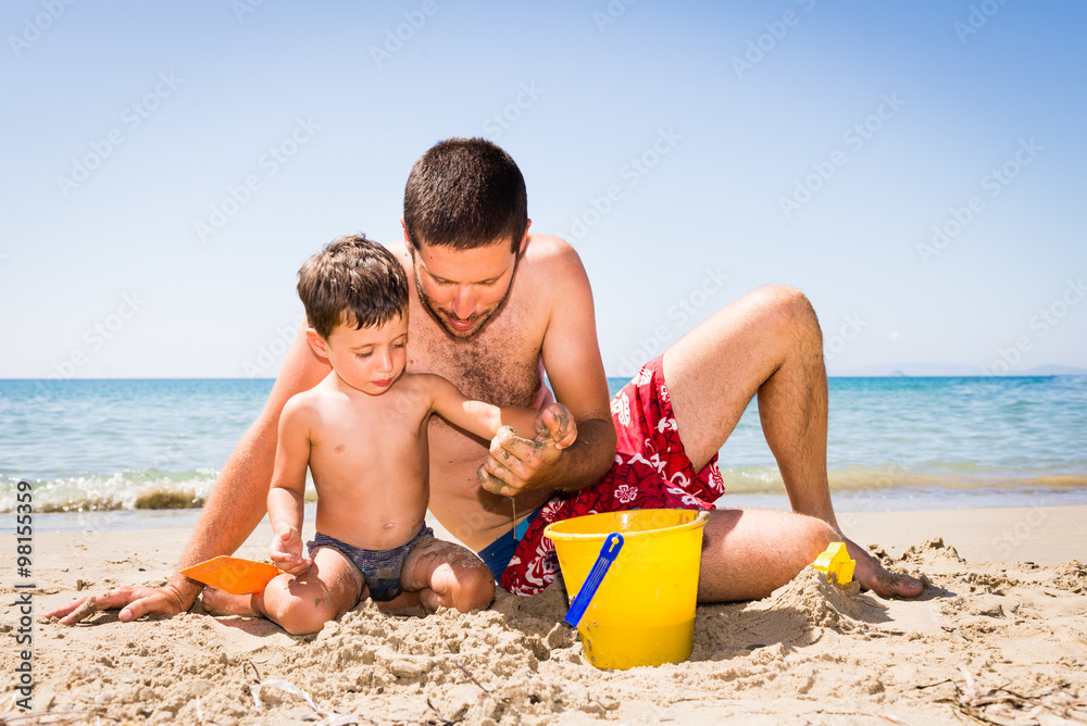 Fototapeta premium close-up of cute and smiling caucasian father and son playing with sand, pail and shovel on the beach with sea background during summer
