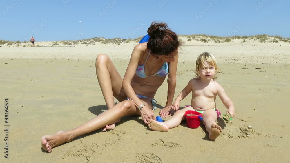 tender family two years age blonde baby with green swimwear with woman mother bikini sitting on beach earth ground playing sand with red bucket and blue and green shovels trying to make a castle tender family two years age blonde baby with green swimwear with woman mother bikini sitting on beach earth ground playing sand with red bucket and blue and green shovels trying to make a castle