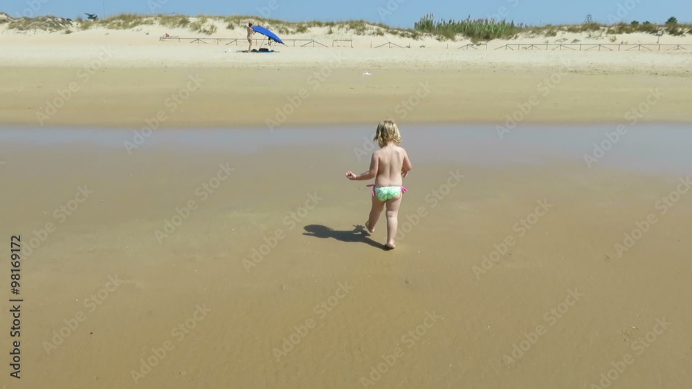 two years blonde baby green swimwear walking, looking for, picking and collecting sea shells in golden sand beach next to woman mother or babysitter two years blonde baby green swimwear walking, looking for, picking and collecting sea shells in golden sand beach next to woman mother or babysitter