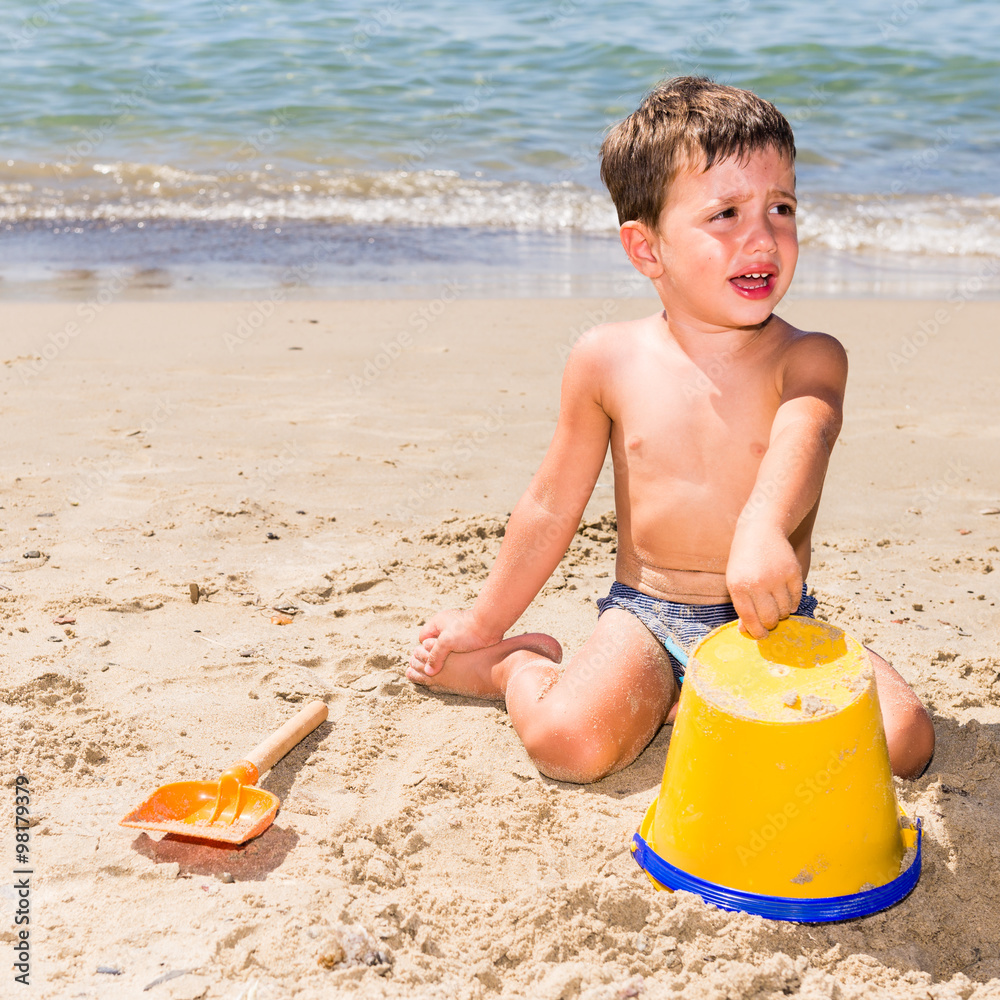 cute caucasian child crying while playing on the beach with pail and shovel in a sunny day with sea background cute caucasian child crying while playing on the beach with pail and shovel in a sunny day with sea background