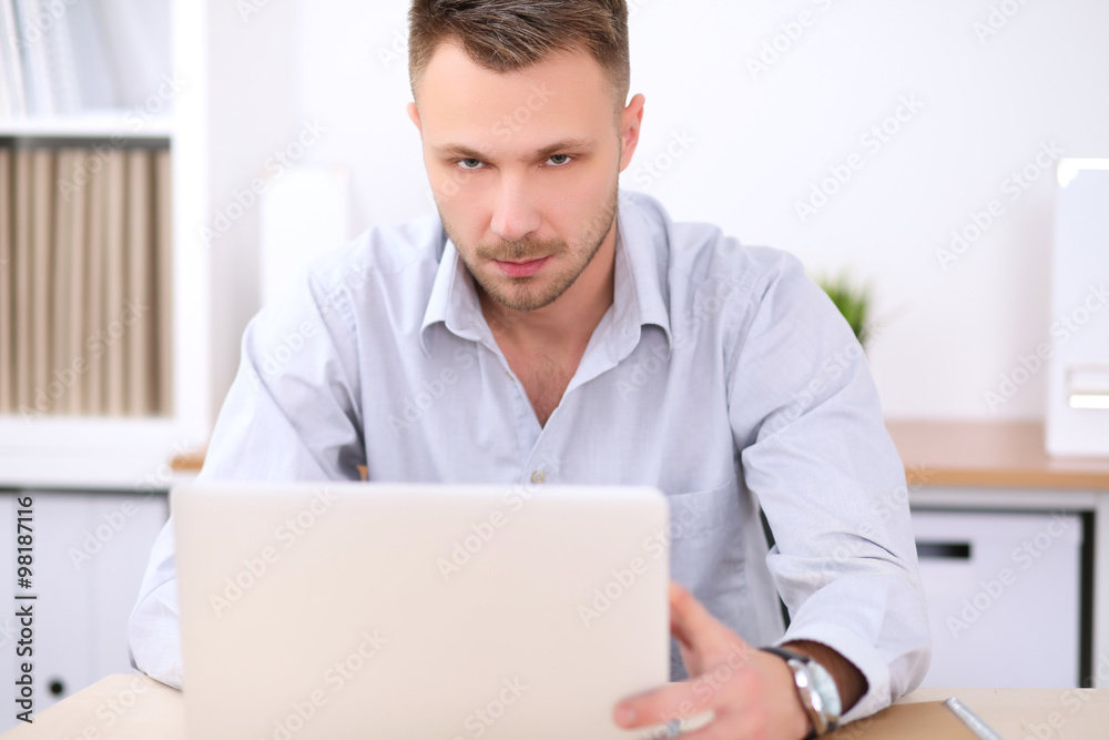 Portrait of young business man sitting at the desk on office background.