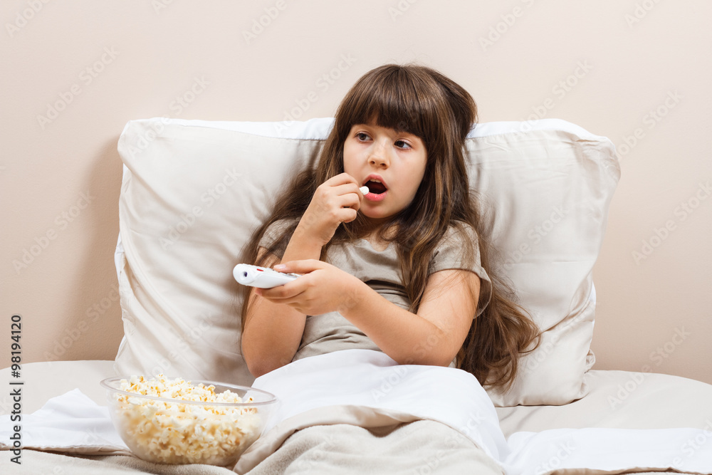 Little girl is watching tv and eating popcorn in her bed. Little girl is watching tv and eating popcorn in her bed.