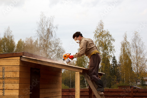 man paints wooden shelter with spray gun