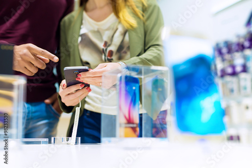 Woman holding mobile phone and black man is pointing finger at phone. They are looking for new smart phone. Mobile shop. Shallow depth of field.