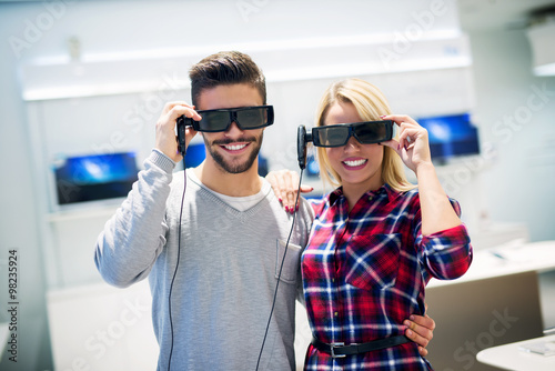 Couple in shopping. They are looking for new 3D tv. They are holding 3D glasses and looking at camera. Shallow depth of field.