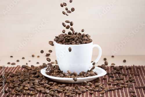 White cup full of brown roasted coffee beans on plate with falling beans from top on tablemat on light wooden textured background.