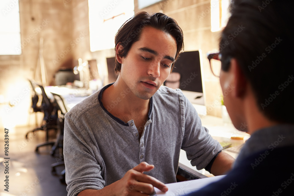 Two Designers Discuss Document During Meeting In Office Two Designers Discuss Document During Meeting In Office