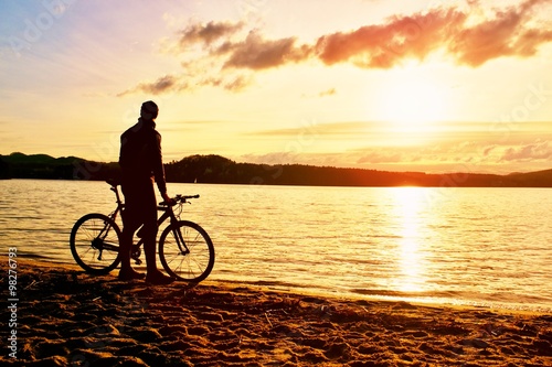 Silhouette of sportsman  holding bicycle on lake beach, colorful  sunset cloudy sky in background and reflection in wavy water level © rdonar
