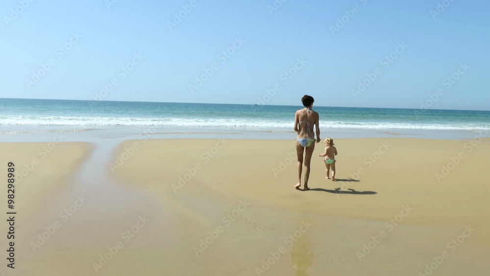 tender family scene of two years age blonde baby with green swimwear with brown hair woman mother blue bikini walking on beach sand seaside with blue ocean and sky tender family scene of two years age blonde baby with green swimwear with brown hair woman mother blue bikini walking on beach sand seaside with blue ocean and sky