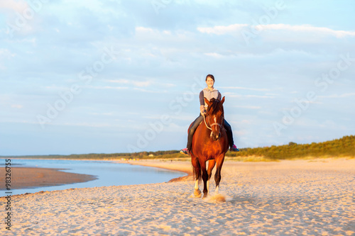 Teenage girl riding horse on the beach at sunset. Outdoors.