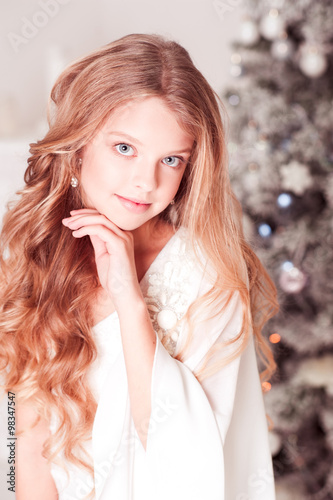 Smiling blonde girl posing over Christmas tree in room. Looking at camera. Teenagerhood. 