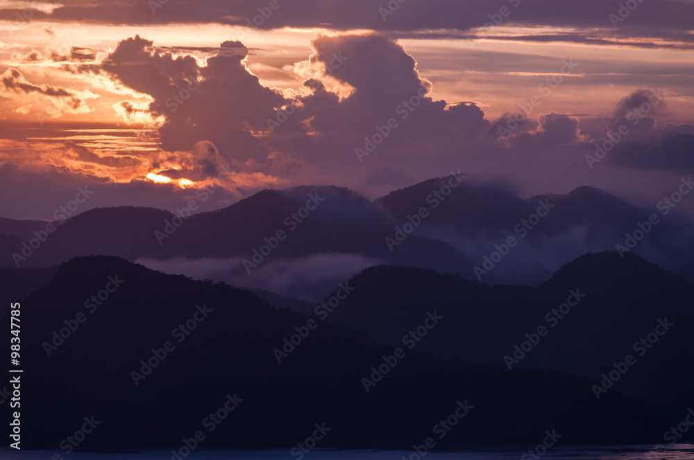 Obraz premium Silhouette of Mountains Above the Water under purple orange cloudscape clouds at sunset
