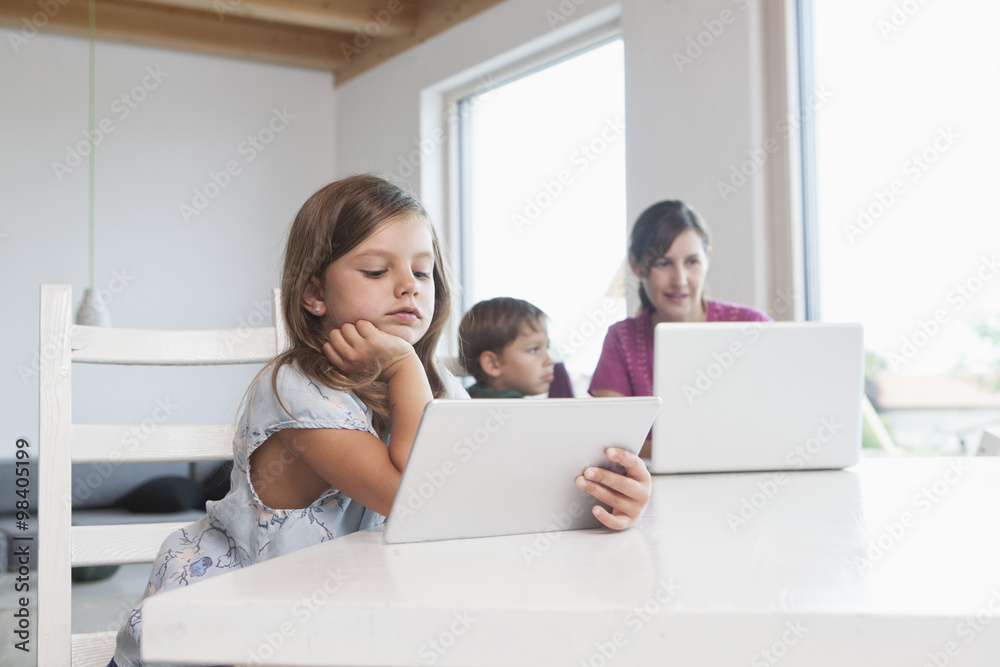 Little girl using digital tablet, mother and brother using laptop in background Little girl using digital tablet, mother and brother using laptop in background