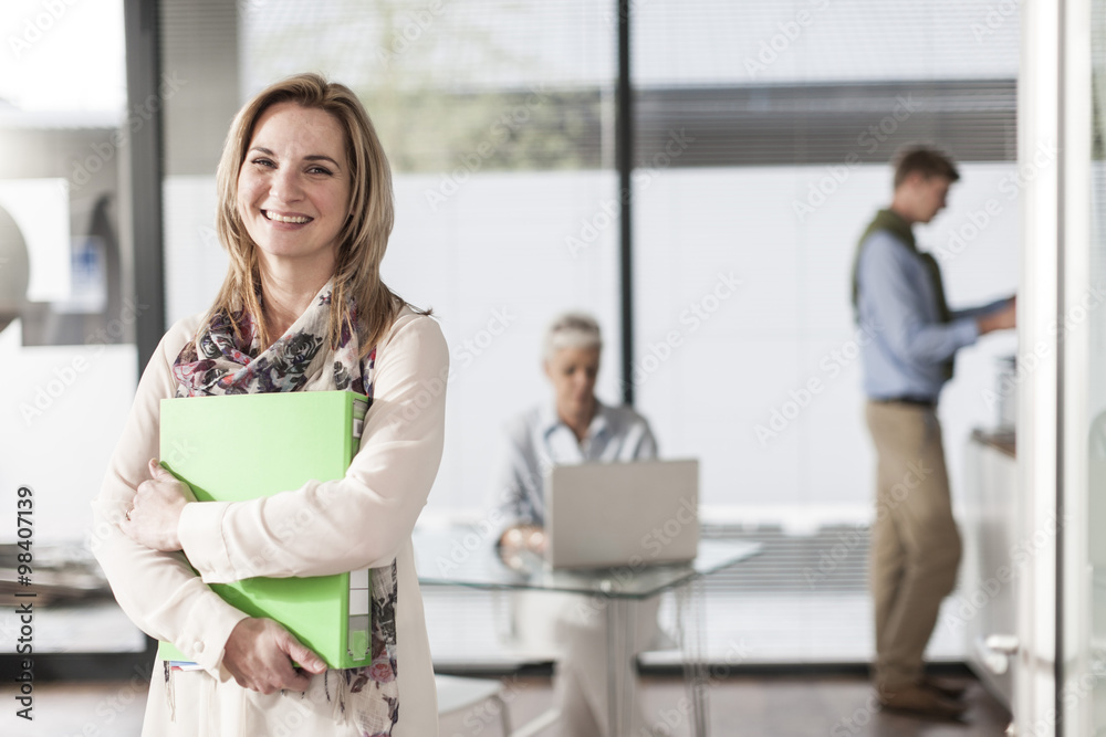 Portrait of smiling woman in office with colleagues in background Portrait of smiling woman in office with colleagues in background