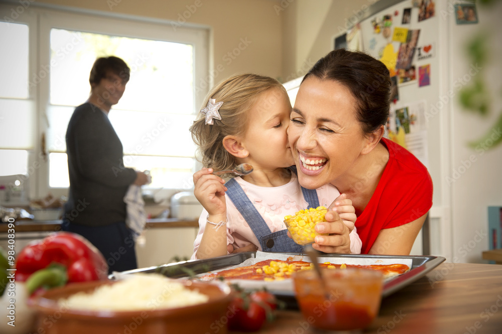 Happy mother and daughter in kitchen preparing pizza with father in background Happy mother and daughter in kitchen preparing pizza with father in background