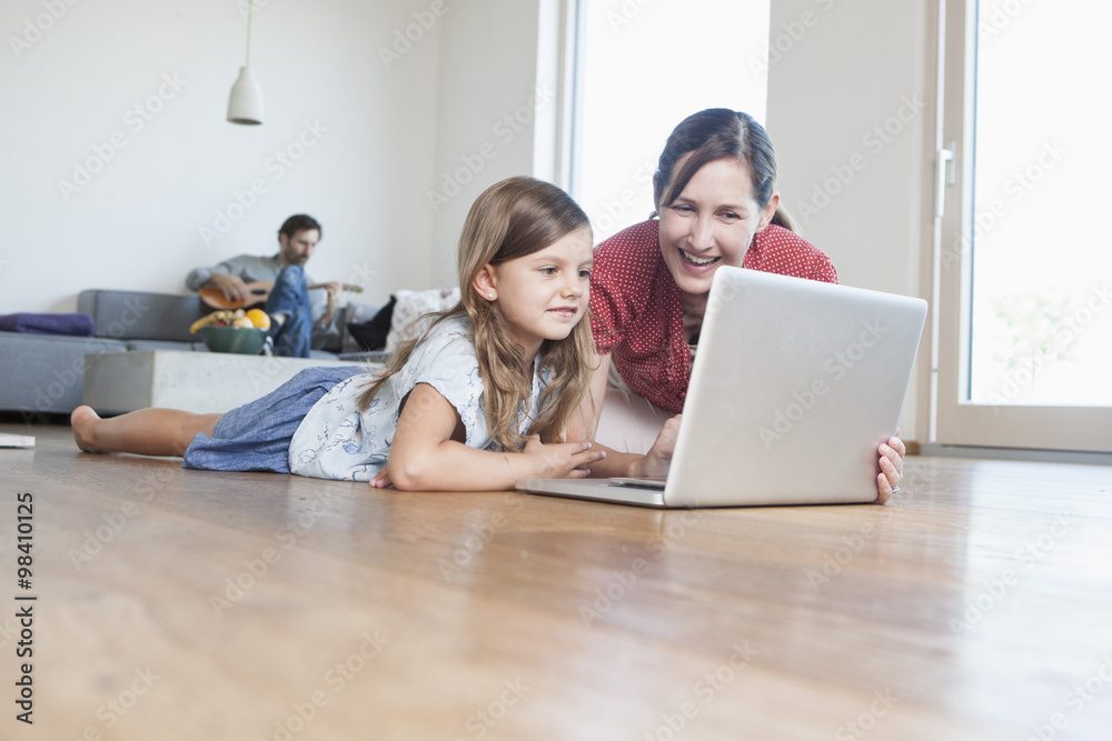 Mother and daughter lying on floor using laptop, father making music in background