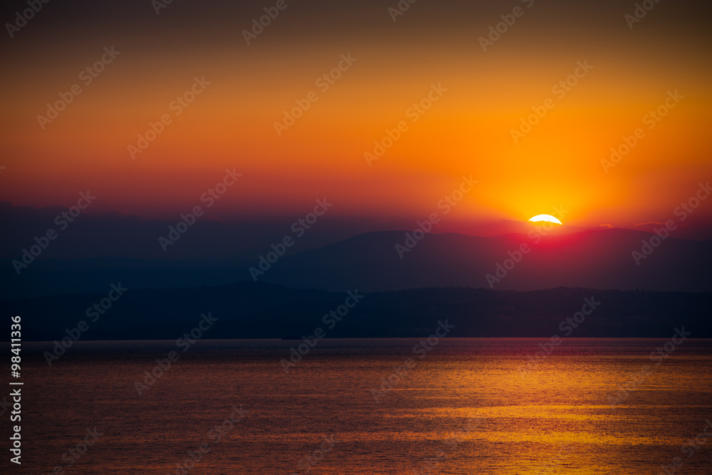 The Rising Sun Over the Sea with Beautiful Vibrant Red Glow. Mountains in Background.