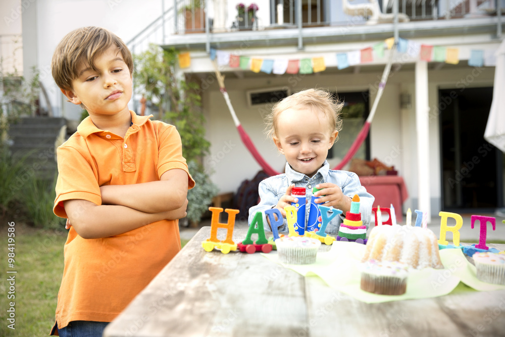 Happy toddler celebrating birthday in garden with displeases brother standing besides Happy toddler celebrating birthday in garden with displeases brother standing besides