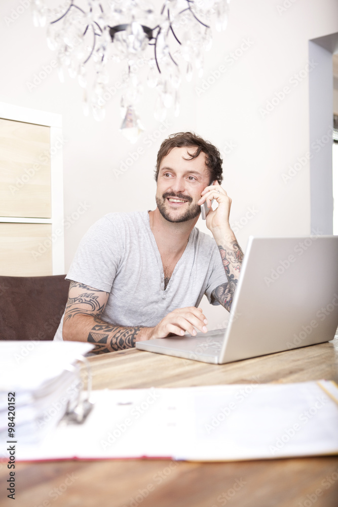 Smiling man sitting at wooden table with laptop and folder telephoning with smartphone Smiling man sitting at wooden table with laptop and folder telephoning with smartphone