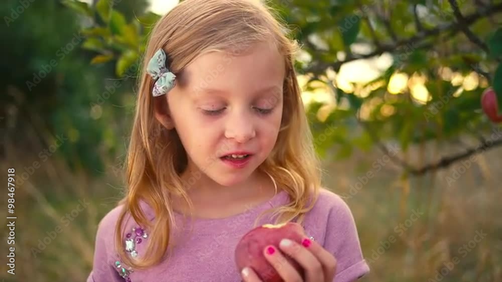 A cute little girl takes a bite out of a fresh apple from a tree and makes a face, and then smiles A cute little girl takes a bite out of a fresh apple from a tree and makes a face, and then smiles