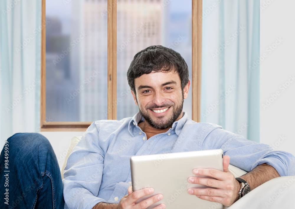 Young attractive Hispanic man at home sitting on white couch using digital tablet Young attractive Hispanic man at home sitting on white couch using digital tablet