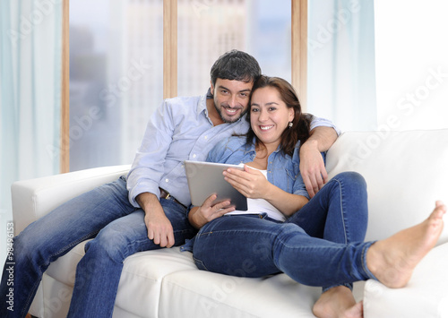 young Latin happy couple on couch at home enjoying using digital tablet