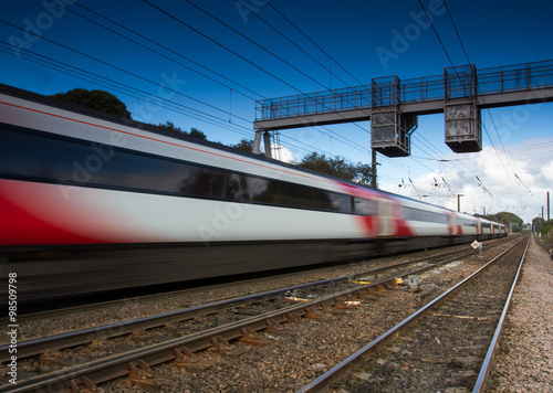 A blurred passenger train speeding past under electric wires.