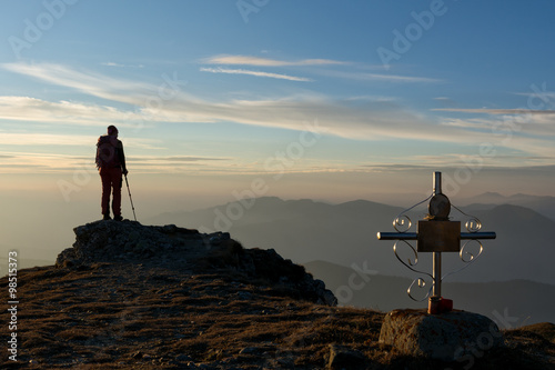 hiker standing on a cliff next to a cross and looking over the horizon at the sunset