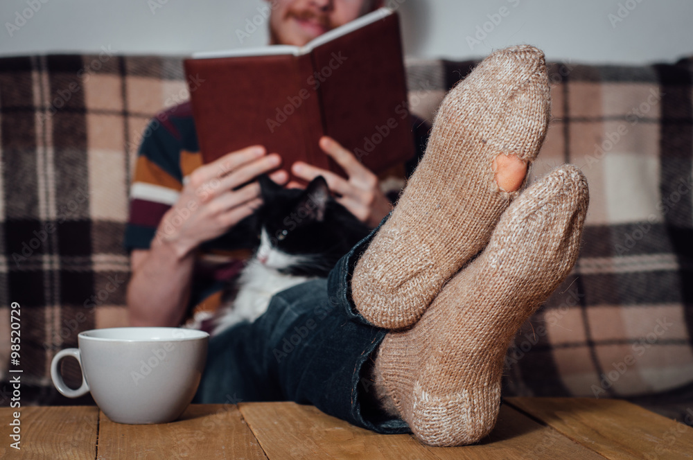 Obraz premium Young man reading book on coach with cat in holey socks