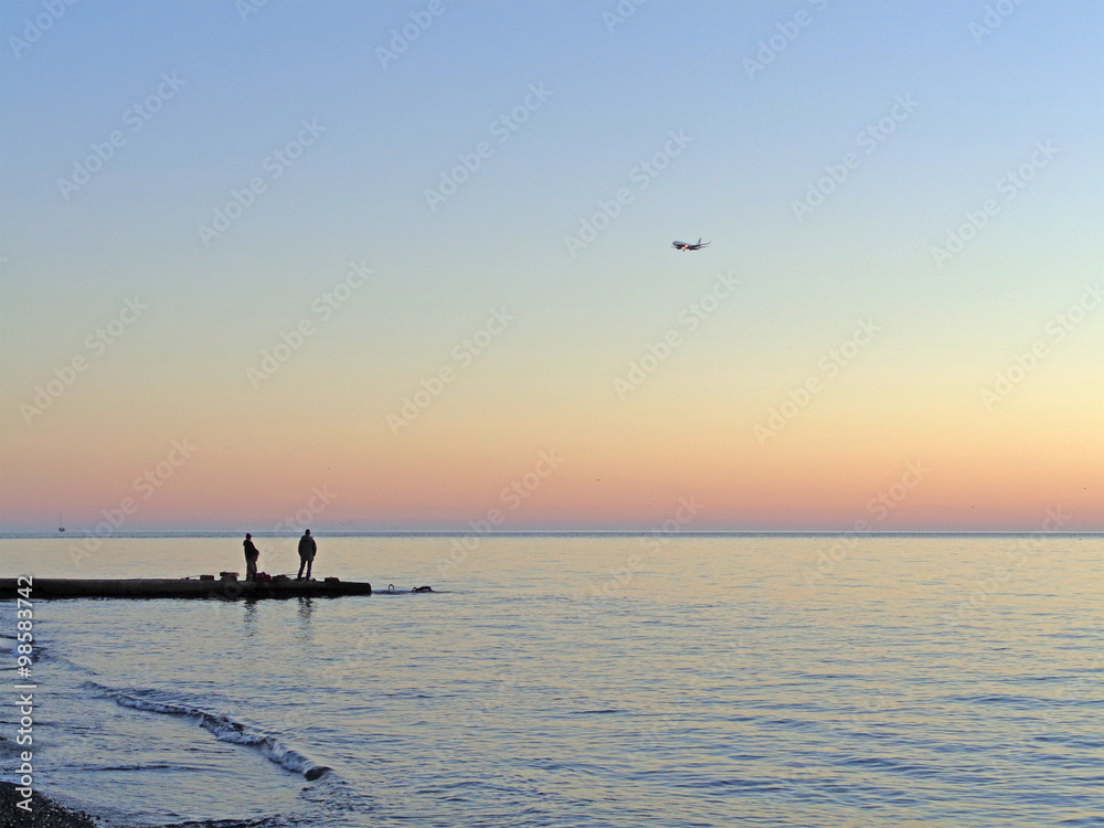The plane flies over the sea and the fishermen on the pier at sunset