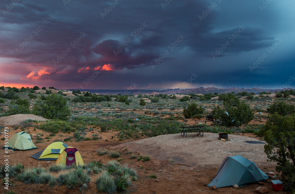 tents in a desert landscape with a dramatic pink and blue thunderstorm sky and arches national park in background
