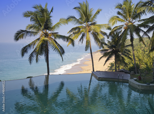 Palm trees are reflected in the pool and the ocean in a background. India.