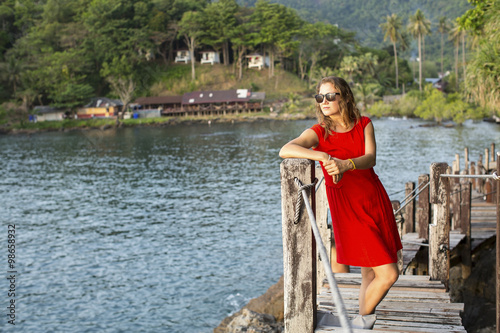 Beautiful girl in a red dress looks at tropical Bay standing on a wooden jetty. © De Visu