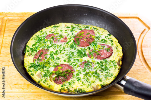 Omlete with sausage and herbs in frying pan on wooden board
