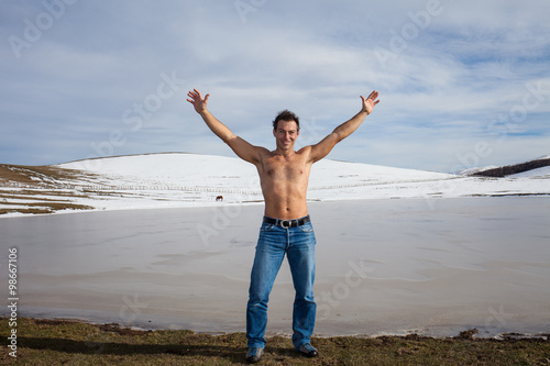 Uomo a torso nudo e braccia alzate in riva a un lago ghiacciato. Sfondo con montagne innevate