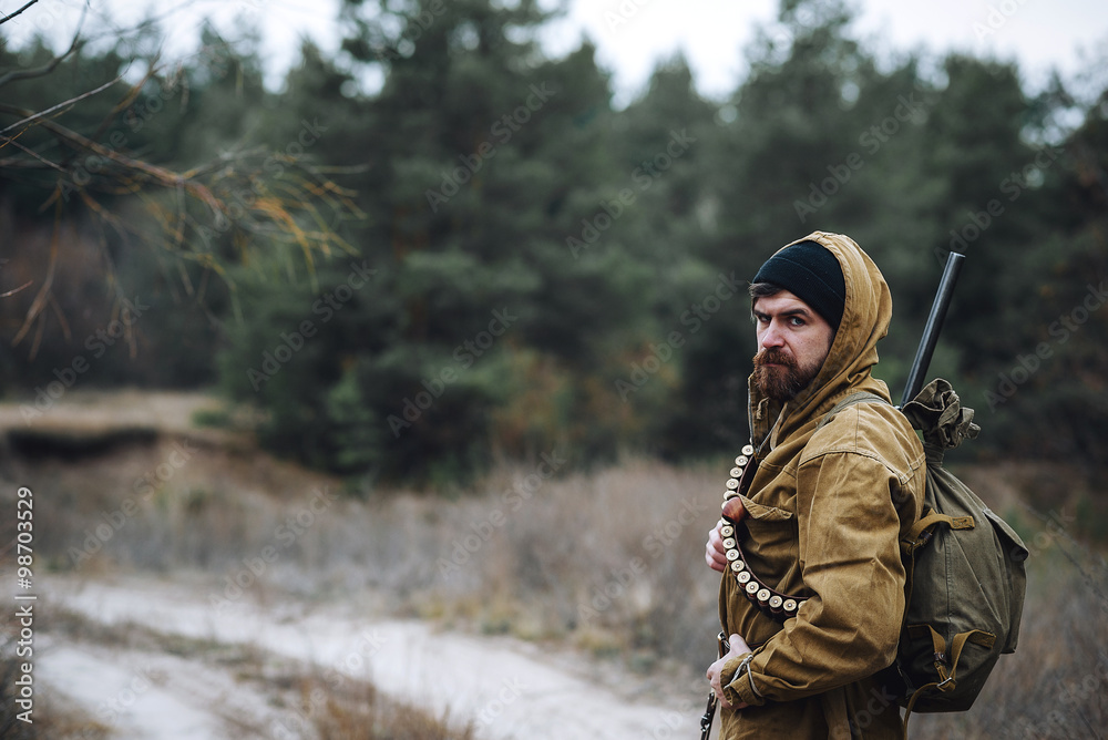brutal hunter, bearded man in warm hat with a gun in his hand, a knife a backpack and smoking pipe in the wild forest in the autumn