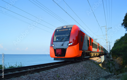 Электричка движется по железнодорожному пути вдоль берега моря в освещении утреннего солнца